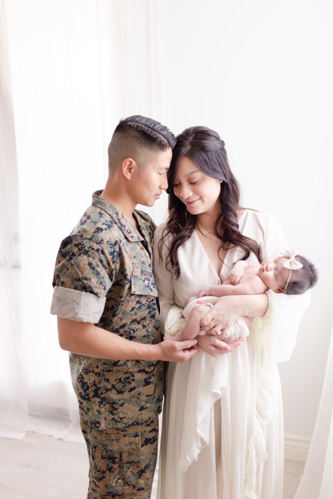 Husband wearing a navy uniform leans into his wife, who is holding their newborn baby girl at their professional newborn photography session in a natural light studio located in southern California.