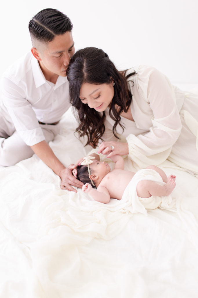 Newborn baby girl lays on white linens and gazes up at her mother, who plays with her hands. Husband leans in and kisses the mother while touching the baby's head.